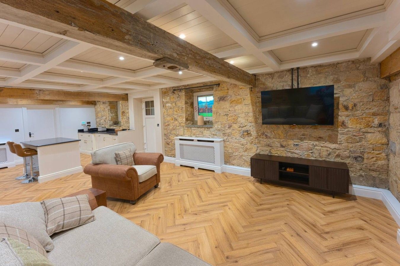 A spacious living area with a stone wall, a flat-screen TV mounted on the wall, a wooden herringbone floor, and a cozy seating area with a sofa and chairs.