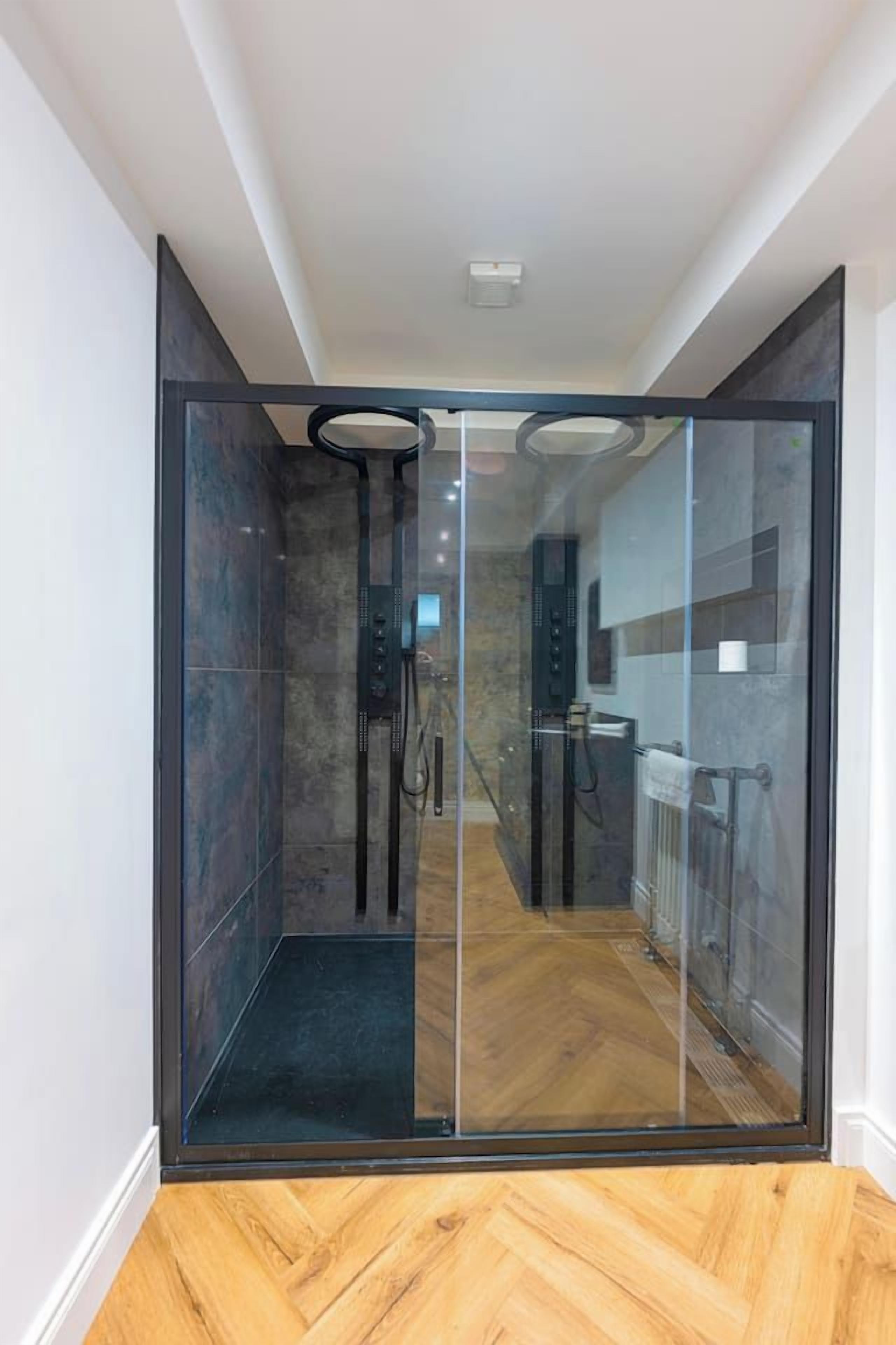 A modern bathroom with a glass shower enclosure, a wooden floor, and a visible shower control panel.