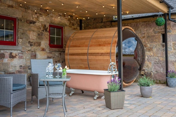 A rustic outdoor setting with a stone wall, a wooden barrel, wicker furniture, and a bathtub. - Distillers Louge by Old Carrick Mill (Carrickmacross)
