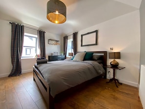 Ein Schlafzimmer mit einem großen Bett, einem Nachttisch mit Lampe, einem Fenster mit Vorhängen und einem an der Wand hängenden Bilderrahmen.