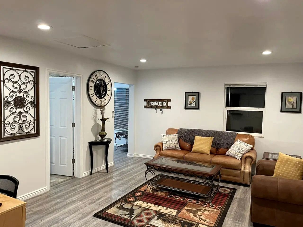 A living room with a brown leather sofa, a coffee table, and a patterned rug. There is a clock on the wall and a 'WELCOME' sign.