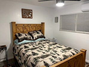 A bedroom with a wooden bed, a ceiling fan, a wall-mounted air conditioning unit, and a window with blinds. - Charming Vacation Rental with Pool & Pet-Friendly Stay in Kanab, Utah (Kanab)