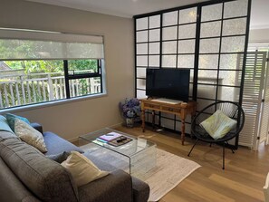 A living room with a sofa, a coffee table, a TV on a wooden stand, a chair, and a large window with a view of greenery. - Seabatical Retreat minutes from the beach (Auckland)