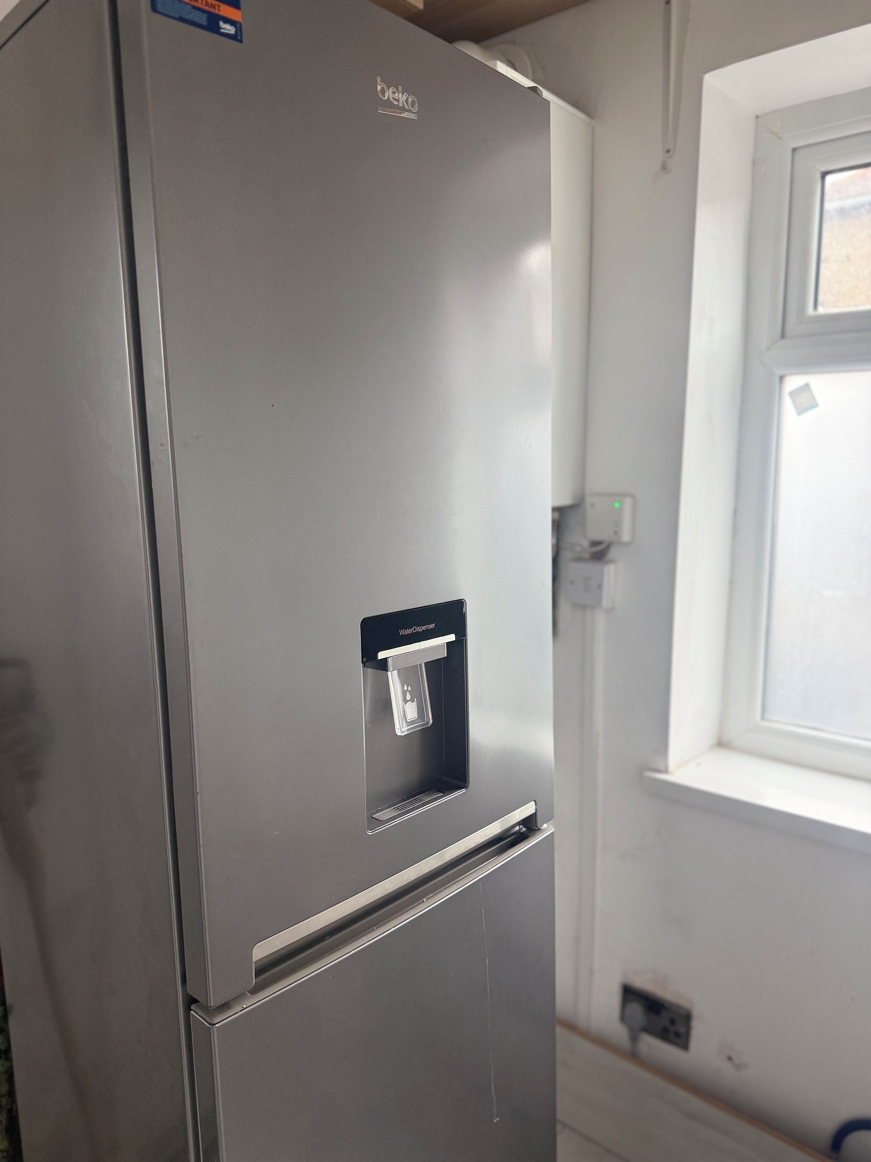 A stainless steel refrigerator with a water dispenser, a window to the right, and a light switch on the wall.