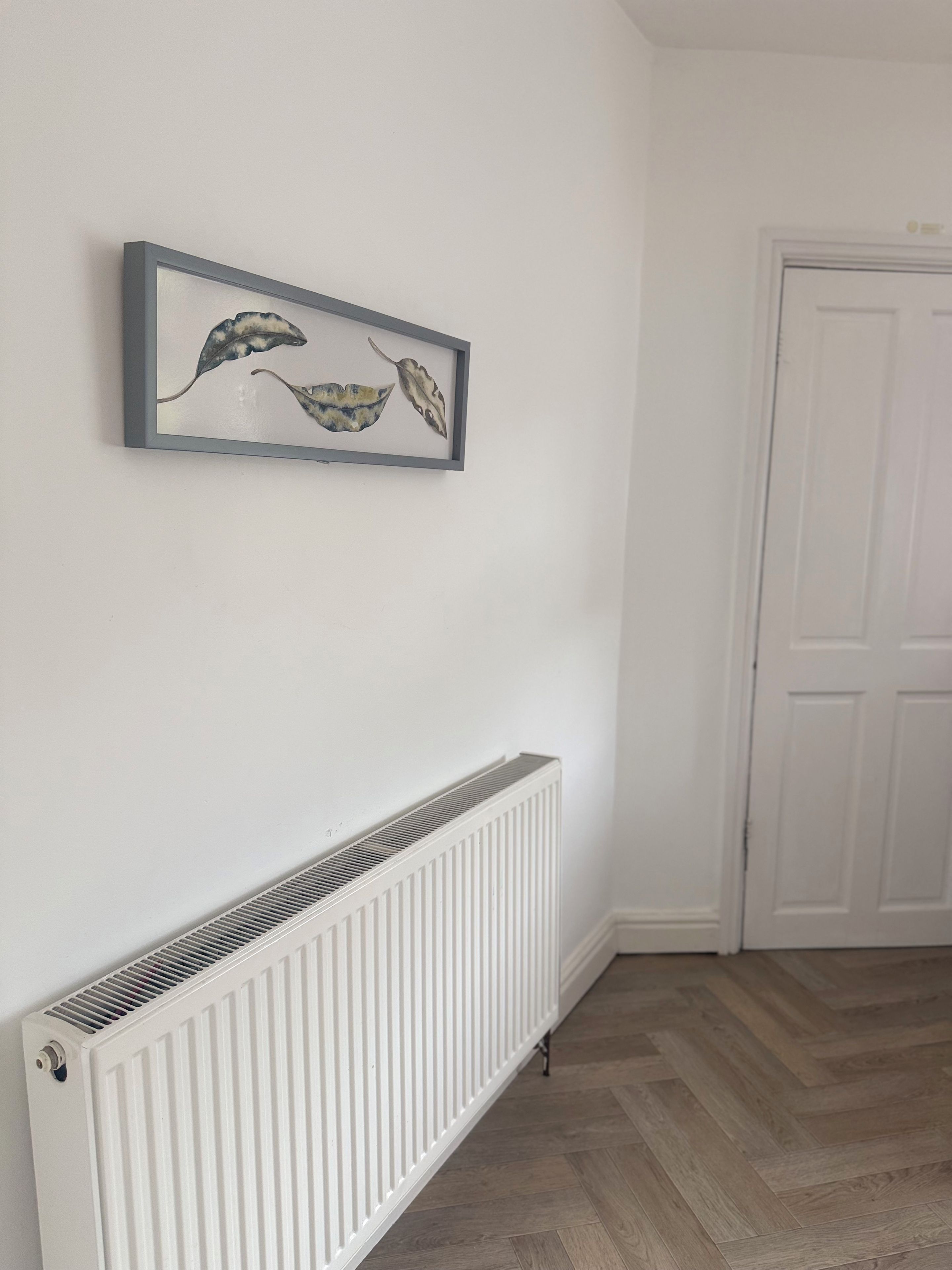 Une pièce avec un radiateur blanc, une œuvre d’art encadrée accrochée au mur et une porte visible au fond.