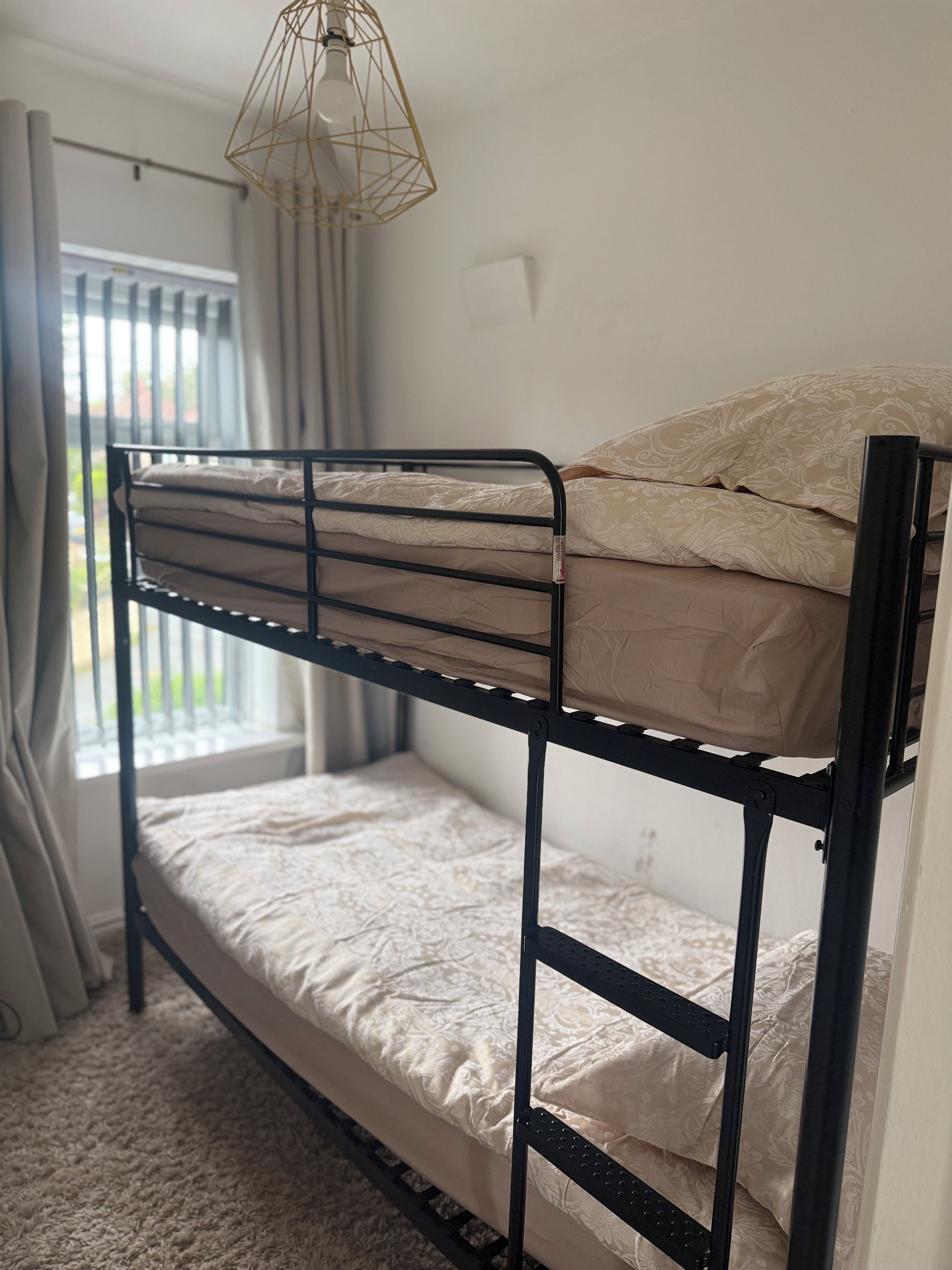 Una cama litera con estructura metálica, una ventana con cortinas y una lámpara.