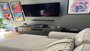Un saló amb un televisor de pantalla plana penjat a la paret, un sofà gris amb un coixí estampat i una petita planta sobre un moble de fusta.