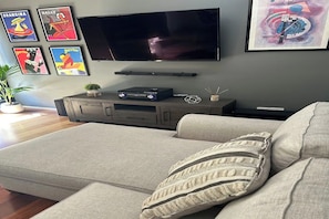 Una sala de estar con un televisor de pantalla plana montado en la pared, un sofá gris con un cojín estampado y una planta pequeña sobre un mueble de madera.