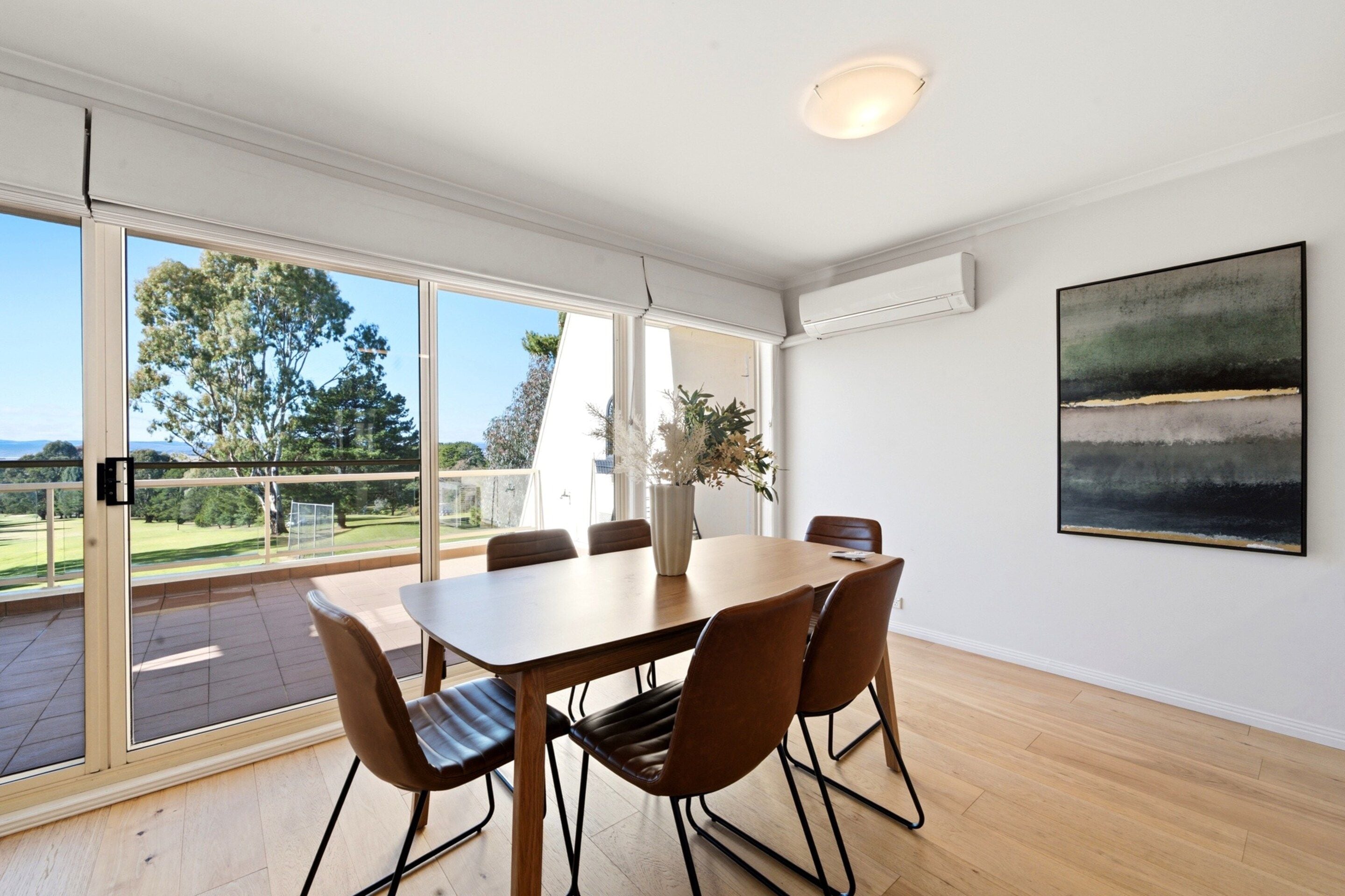 A modern dining room with a large table and chairs, large windows with a view of greenery, and an air conditioning unit on the wall.