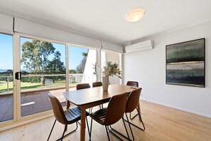 A modern dining room with a large table and chairs, large windows with a view of greenery, and an air conditioning unit on the wall. - Spacious 2-bedroom Retreat With Golf Course Views (Canberra)