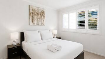A bedroom with a bed, two bedside tables, a wall-mounted artwork, and a window with blinds.