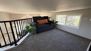 Une pièce avec un tapis gris, un canapé noir orné d’un coussin rouge et une fenêtre donnant sur des arbres.