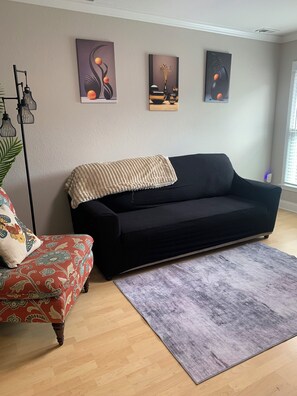 Een woonkamer met een zwarte bank, een fauteuil met een patroon en een grijs vloerkleed. Boven de bank hangen drie kunstwerken aan de muur.