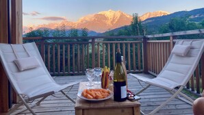 A wooden deck with two lounge chairs, a table with a bottle of wine, glasses, and a plate of fruit. - Vrbo Property (Combloux)