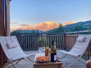 A wooden deck with two lounge chairs, a table with a bottle of wine, glasses, and a plate of fruit. - Vrbo Property (Combloux)