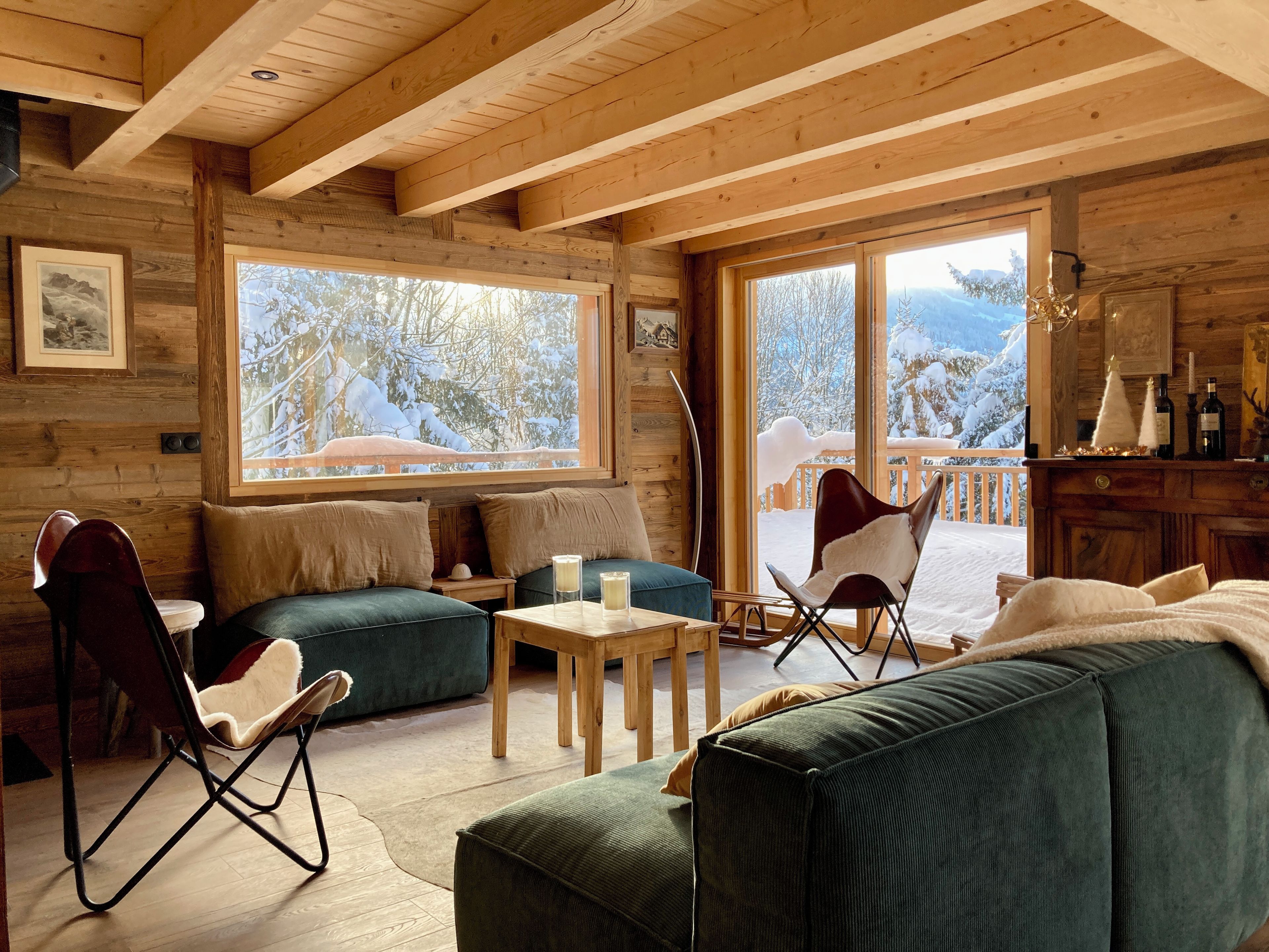 A cozy cabin interior with wooden walls, a sofa, a chair, a table, and a large window offering a snowy mountain view.