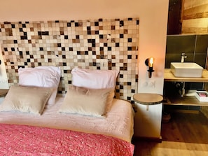 A bedroom with a checkered headboard, a bed with two pillows, a bedside table with a lamp, and a bathroom with a sink and mirror. - Vrbo Property (Combloux)