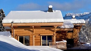 A wooden chalet with a snow-covered roof, large windows, and a balcony overlooking a snowy landscape with mountains in the background. - Vrbo Property (Combloux)