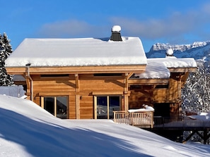 A wooden chalet with a snow-covered roof, large windows, and a balcony overlooking a snowy landscape with mountains in the background. - Vrbo Property (Combloux)