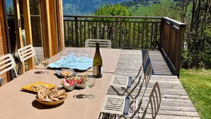 An outdoor dining area with a table set for four, a bottle of wine, and a view of snow-capped mountains. - Vrbo Property (Combloux)