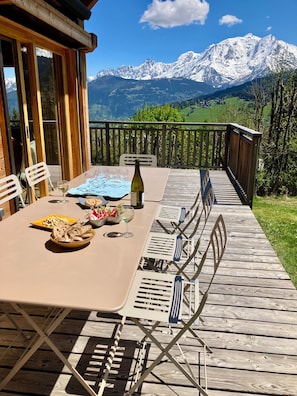 An outdoor dining area with a table set for four, a bottle of wine, and a view of snow-capped mountains. - Vrbo Property (Combloux)