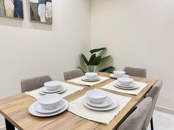 A dining table set with white plates and bowls, surrounded by chairs, in a room with a plant and artwork on the wall. - Sahab - Pearl Tower 1BHK 5 min From Alkhan Beach (Sharjah)