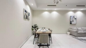 Et moderne spiseområde med et rektangulært bord dækket til fire personer, omgivet af stole, i et rum med hvide gulv og vægge, en loftlampe og en sofa i baggrunden.