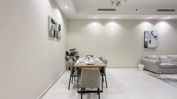 A modern dining area with a rectangular table set for four, surrounded by chairs, in a room with a white floor and walls, a ceiling light fixture, and a sofa in the background.