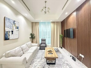 A modern living room with a sofa, armchair, coffee table, and a flat-screen TV mounted on the wall. - Sahab - Pearl Tower 1BHK 5 min From Alkhan Beach (Sharjah)