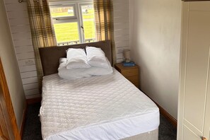 A neatly made bed with a quilted cover, a bedside table with a lamp, and a window with curtains. - Cabin Sleeps 2 With Heating (Mablethorpe)