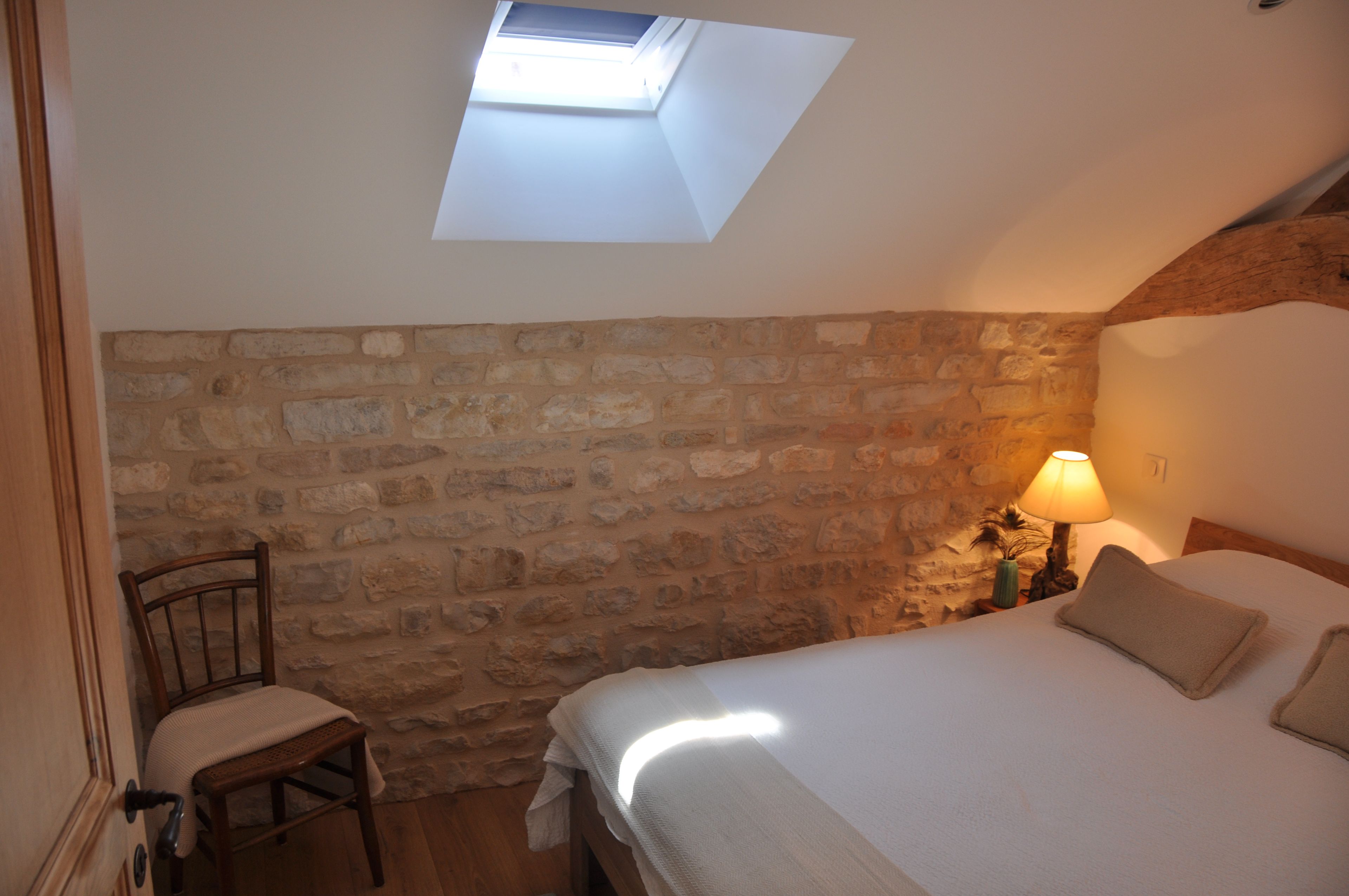 Een slaapkamer met een schuine plafond, een stenen muur, een enkel bed met kussens, een stoel en een nachtkastje met een lamp.