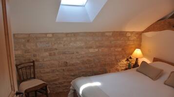 Een slaapkamer met een schuine plafond, een stenen muur, een enkel bed met kussens, een stoel en een nachtkastje met een lamp.
