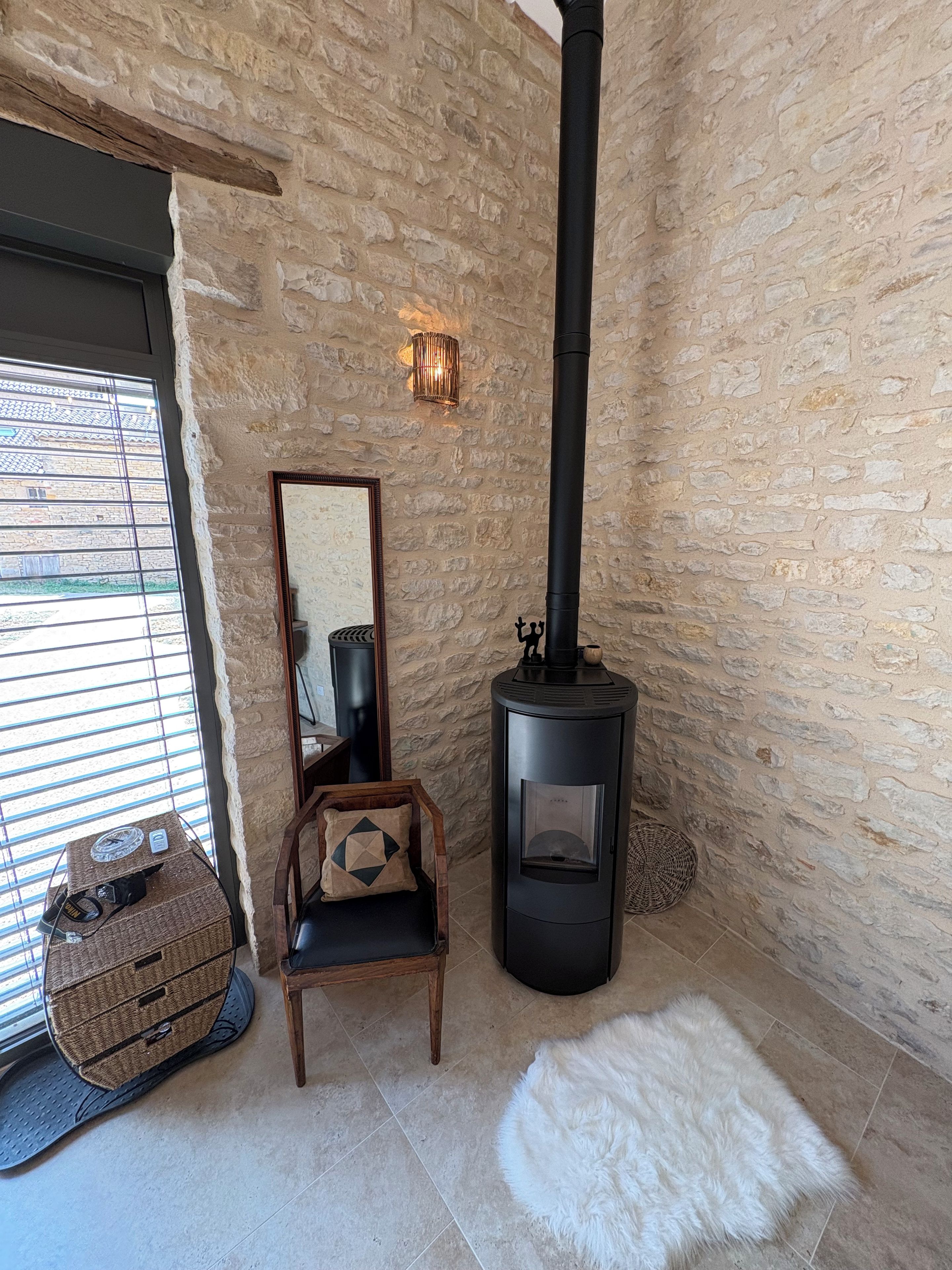 A stone-walled room with a black wood-burning stove, a wooden chair, a wicker basket, and a mirror on the wall.