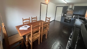 Un comedor con mesa y sillas de madera, una lámpara colgante y vista a una cocina con electrodomésticos modernos.