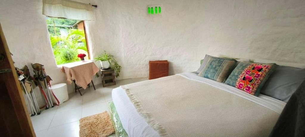 Ein Schlafzimmer mit einem Bett, Kissen, einem kleinen Tisch mit einer Pflanze und einem Fenster mit Blick auf Grün.