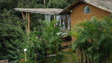 Uma cabana rústica de madeira com telhado de palha, cercada por vegetação exuberante e um deck de madeira.