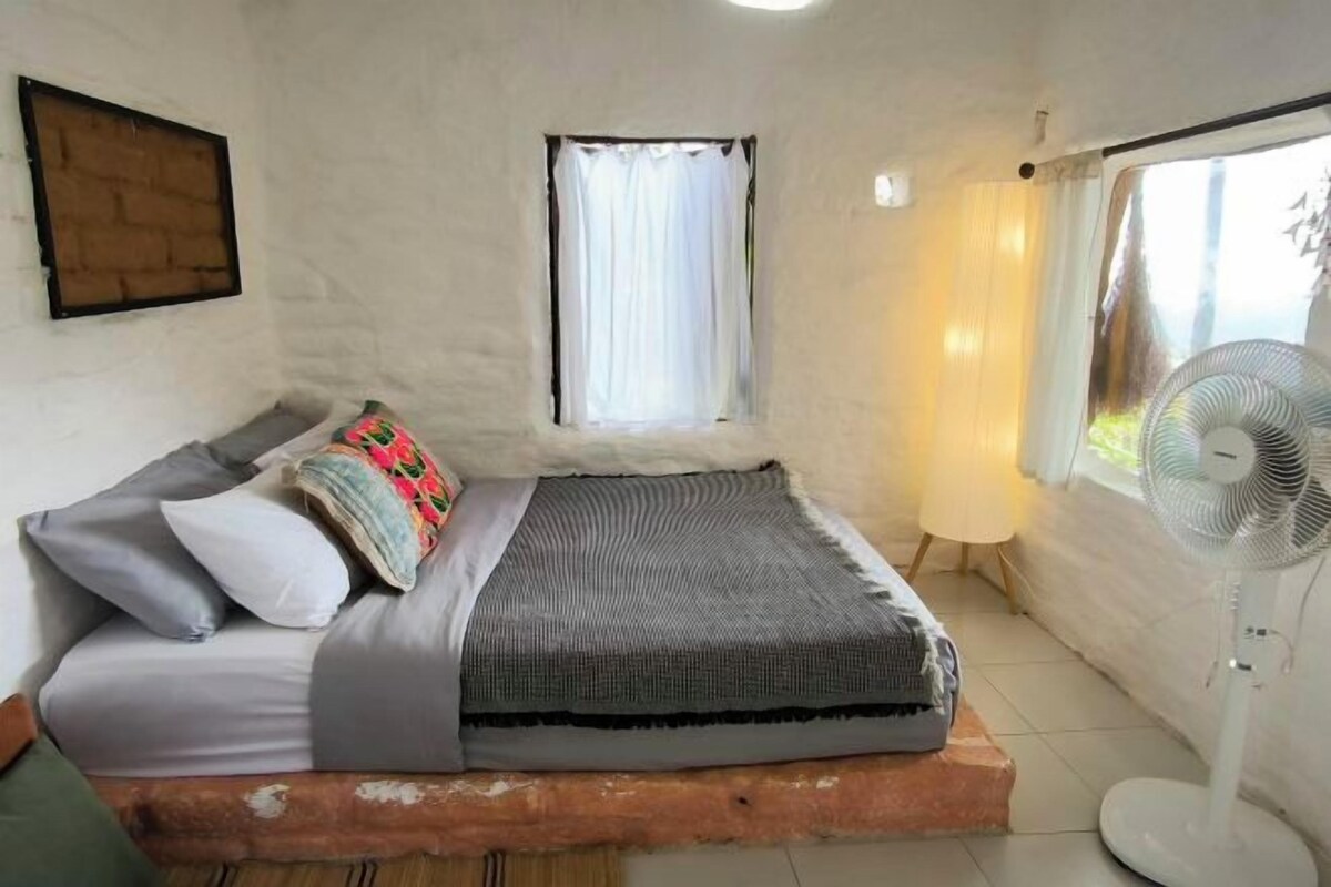A single bed with a wooden headboard, a bedside table, a window with curtains, and a standing fan.