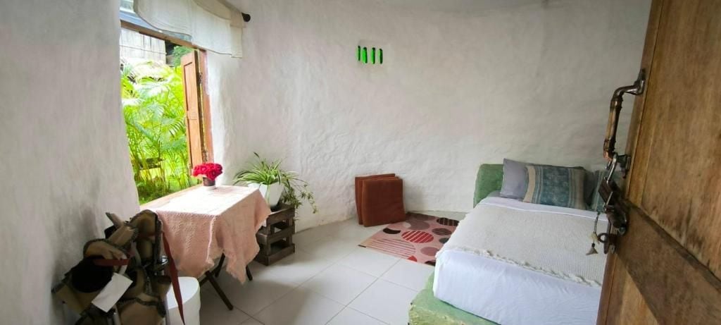 A small, cozy room with a bed, a table with a plant, and a window with a view of greenery.