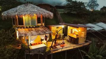 A thatched-roof structure with a wooden deck and a small attached building, surrounded by vegetation.