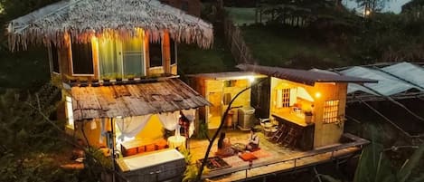 Estrutura com telhado de palha, deck de madeira e um pequeno edifício anexo, tudo cercado por vegetação.
