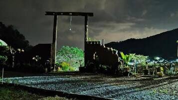 A nighttime view of a rural area with a wooden structure, possibly a gate or entrance, surrounded by greenery and plants.
