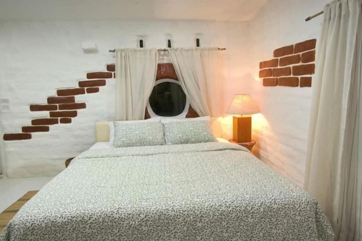 A bedroom with a bed, a window with curtains, a brick wall, and a nightstand with a lamp.