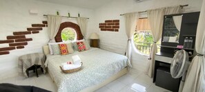 Un dormitorio con cama, una mesita, una silla, un ventilador y una ventana con cortinas.
