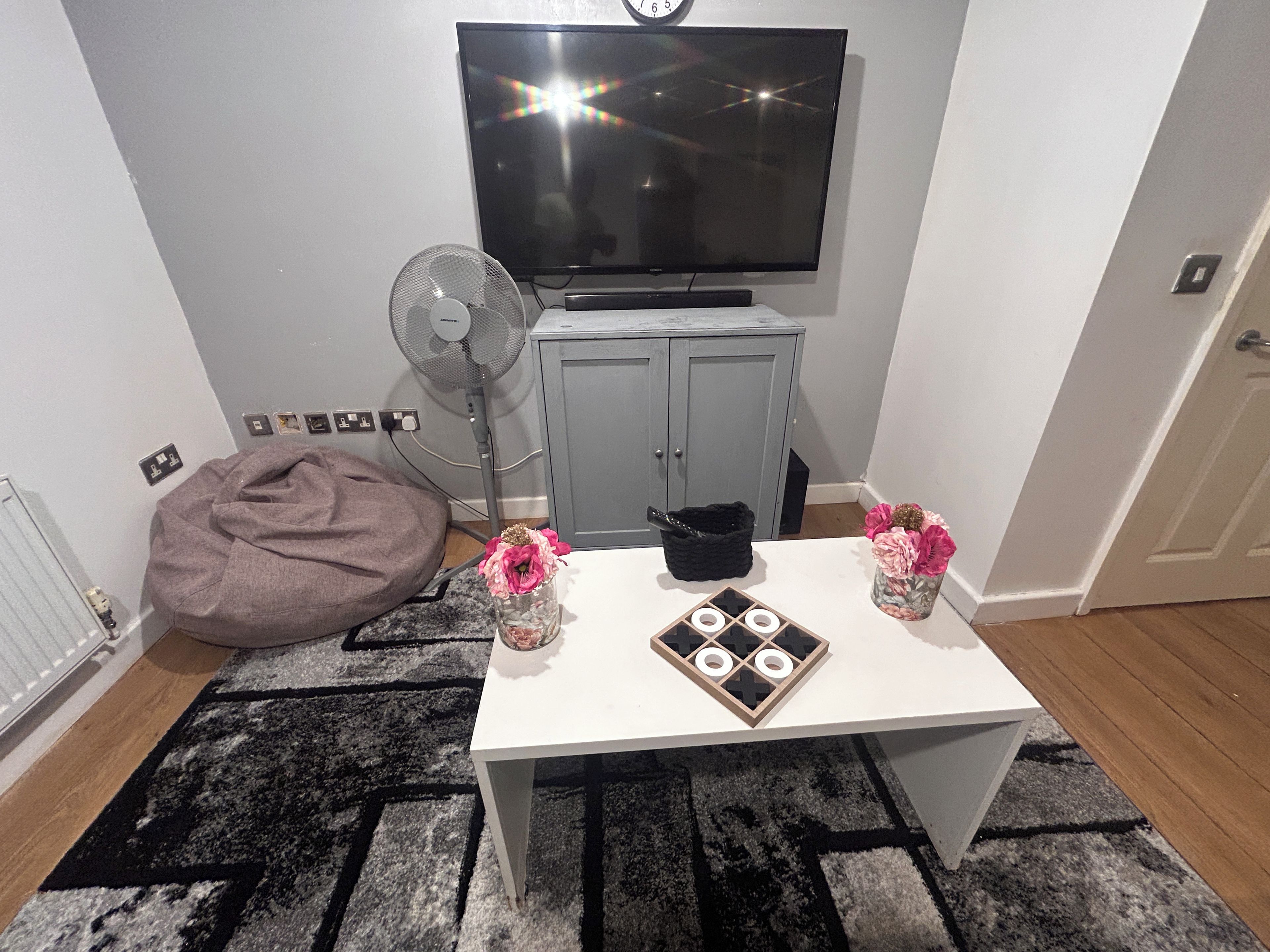 Ett rum med en tv, en fläkt, ett bord med dekorativa föremål, en sittpuff och ett skåp.