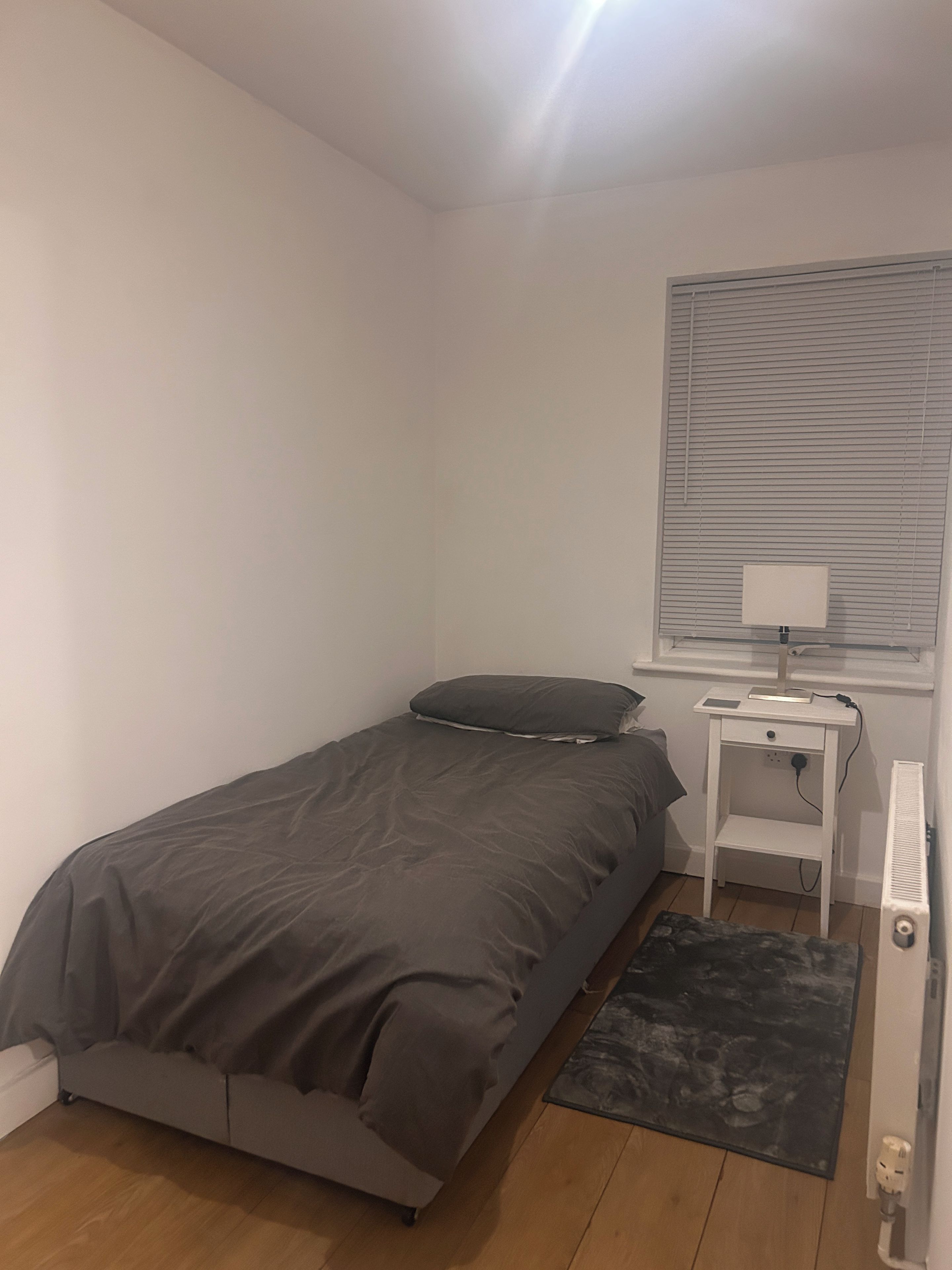 A small, single-bed room with a grey bedspread, a bedside table with a lamp, a radiator, and a window with blinds.