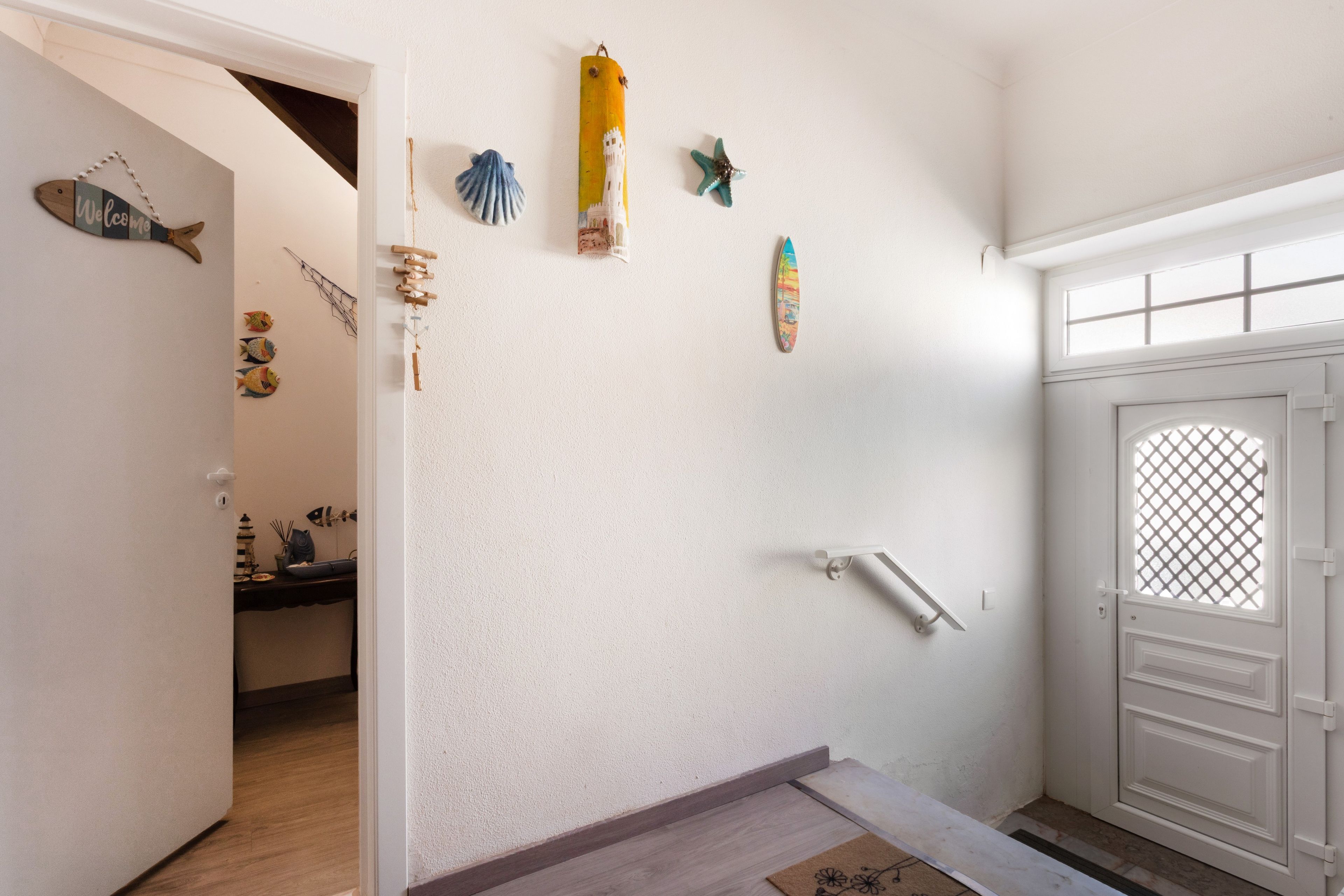 Un pasillo con una puerta blanca, una ventana pequeña y diversas decoraciones en las paredes, incluyendo una tabla de surf y peces.