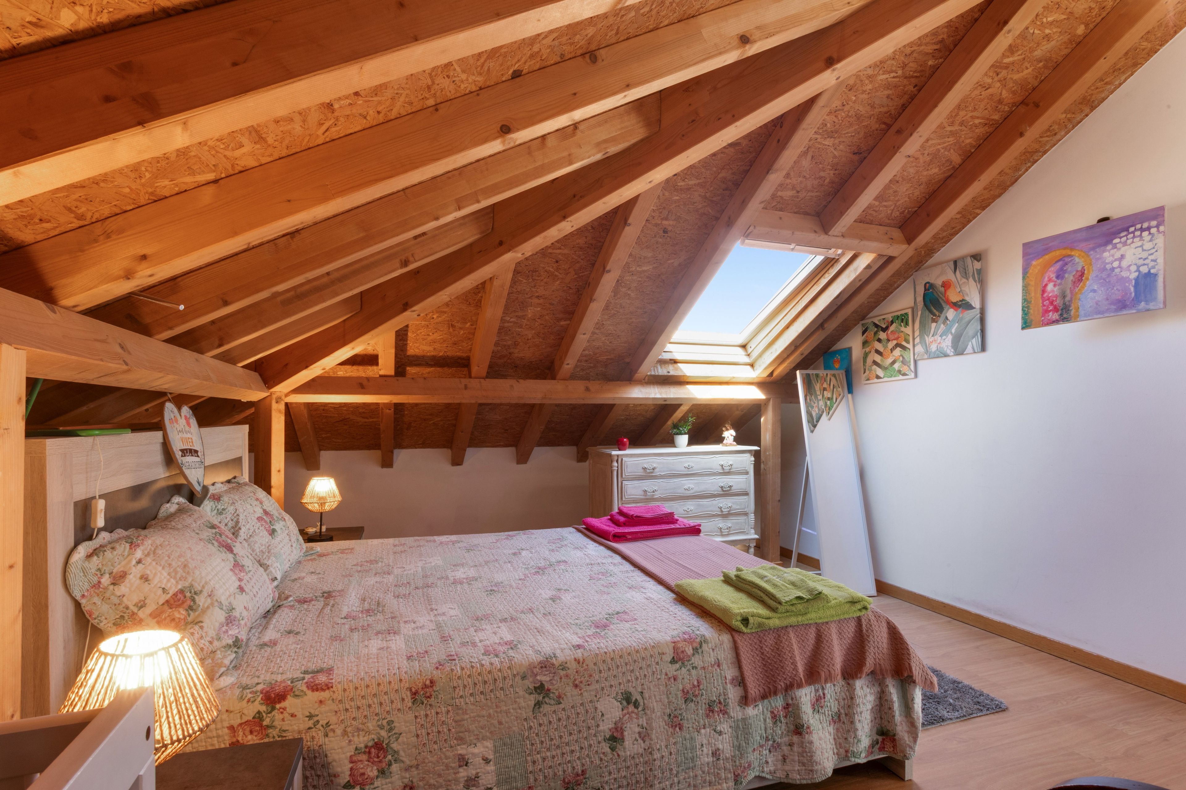 Un dormitorio con techo inclinado, vigas de madera, tragaluces, cama con ropa de cama floral, cómoda y decoración en la pared.