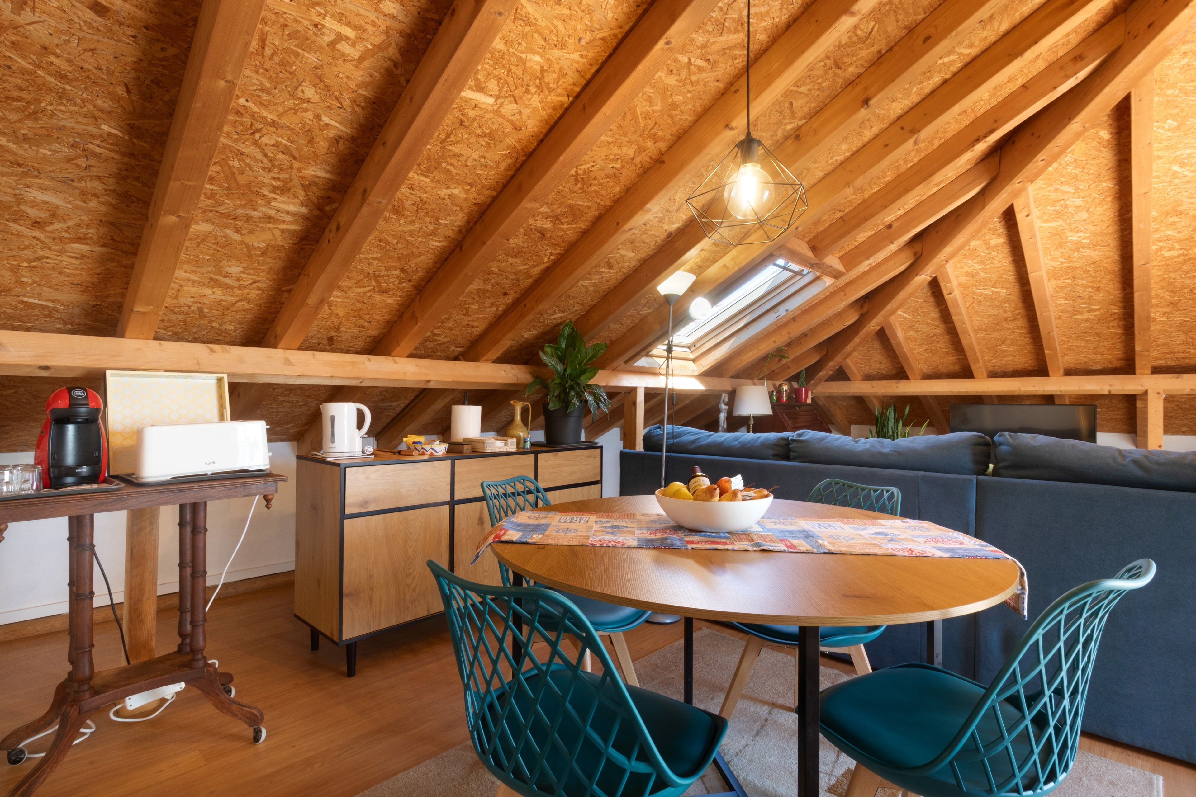 Una habitación acogedora en el desván, con techo de madera, una mesa de comedor con sillas, un sofá y una zona de cocina con electrodomésticos.