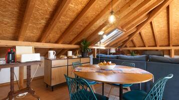 Una habitación acogedora en el desván, con techo de madera, una mesa de comedor con sillas, un sofá y una zona de cocina con electrodomésticos.