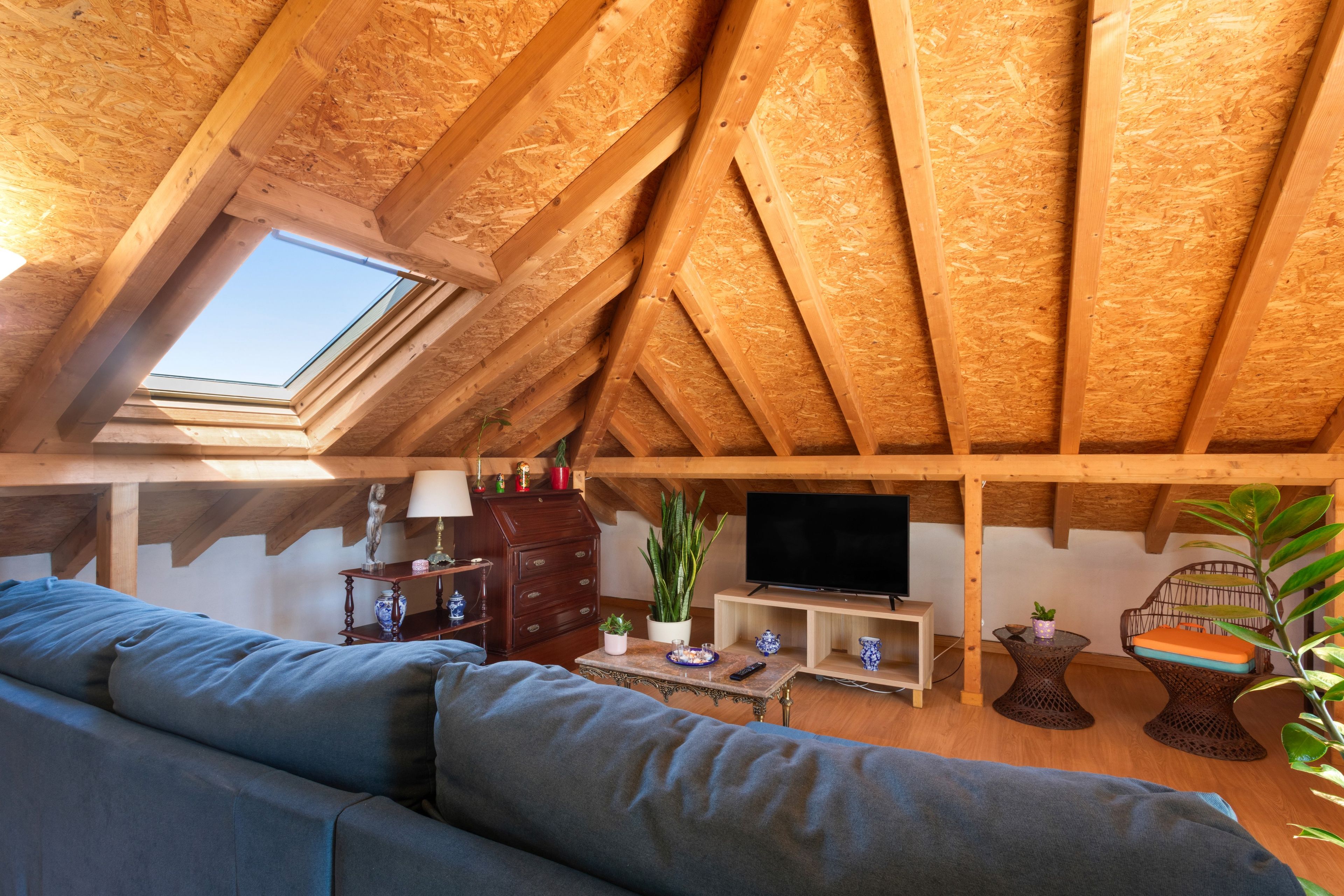 Quarto no sótão com teto inclinado, claraboia, televisão, sofá e diversos objetos decorativos.